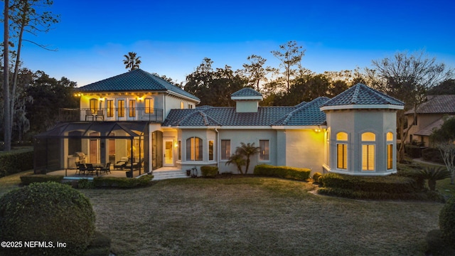 back house at dusk with a patio, a balcony, and a lawn