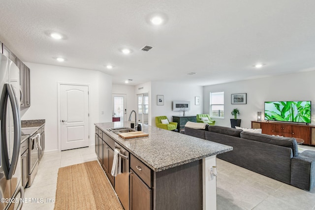 kitchen with sink, light tile patterned floors, appliances with stainless steel finishes, dark brown cabinets, and an island with sink