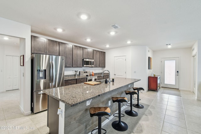 kitchen with dark brown cabinetry, a breakfast bar area, appliances with stainless steel finishes, an island with sink, and dark stone counters