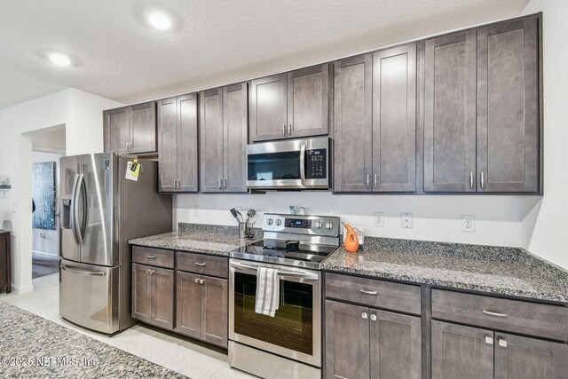 kitchen with light tile patterned floors, appliances with stainless steel finishes, dark stone countertops, dark brown cabinets, and a textured ceiling