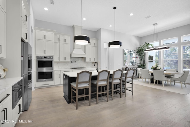 kitchen featuring white cabinetry, a center island with sink, decorative light fixtures, tasteful backsplash, and double oven