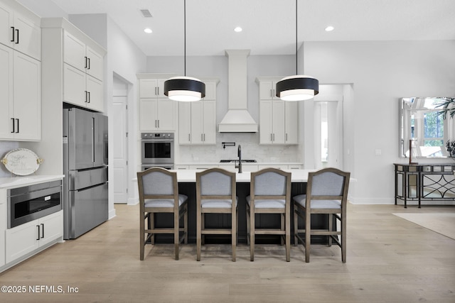 kitchen featuring stainless steel appliances, an island with sink, custom range hood, decorative light fixtures, and white cabinetry