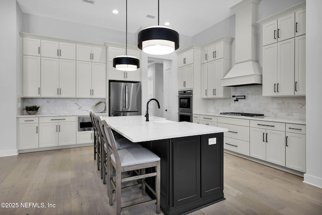 kitchen with white cabinetry, a center island with sink, sink, custom range hood, and appliances with stainless steel finishes