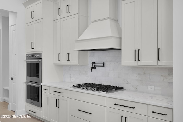 kitchen with appliances with stainless steel finishes, backsplash, premium range hood, white cabinetry, and light stone countertops