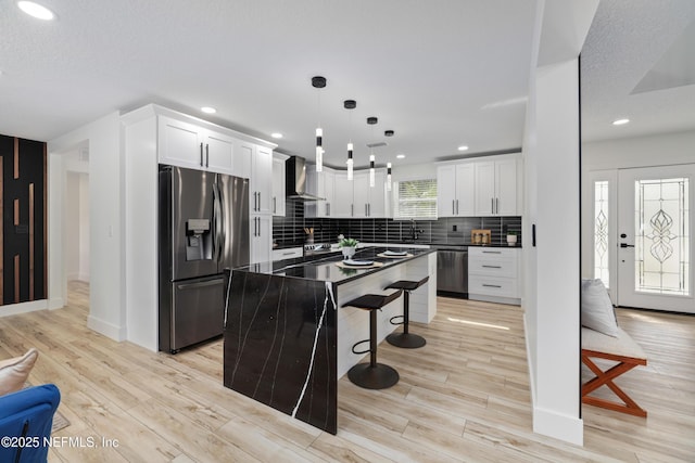 kitchen featuring appliances with stainless steel finishes, white cabinetry, hanging light fixtures, a center island, and wall chimney range hood