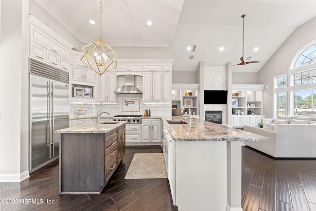 kitchen with wall chimney exhaust hood, sink, built in appliances, decorative light fixtures, and a kitchen island with sink