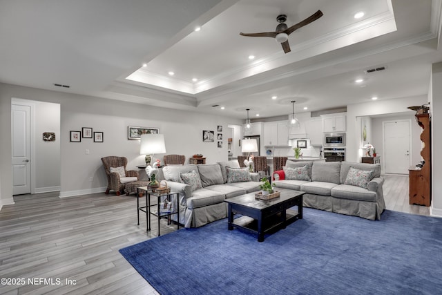 living room with light wood-style floors, a tray ceiling, visible vents, and ornamental molding