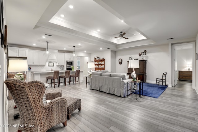 living room with a raised ceiling, a ceiling fan, light wood-style flooring, crown molding, and recessed lighting