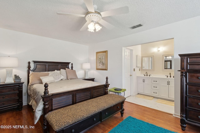 bedroom with visible vents, wood finished floors, a textured ceiling, ensuite bath, and a ceiling fan