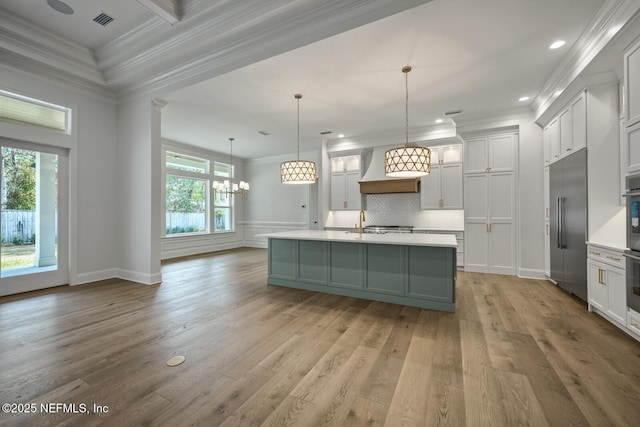 kitchen featuring stainless steel built in refrigerator, custom exhaust hood, decorative light fixtures, ornamental molding, and an island with sink