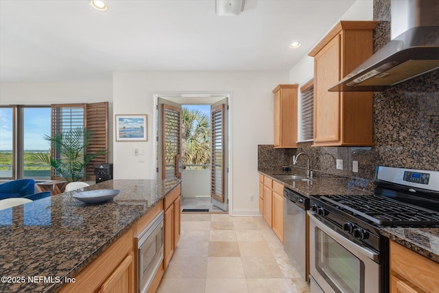 kitchen with wall chimney range hood, sink, dark stone counters, and appliances with stainless steel finishes