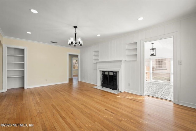 unfurnished living room featuring built in shelves, ornamental molding, a chandelier, and light hardwood / wood-style flooring