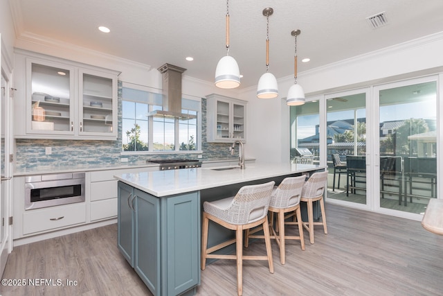 kitchen featuring decorative light fixtures, backsplash, island exhaust hood, light hardwood / wood-style floors, and a center island with sink