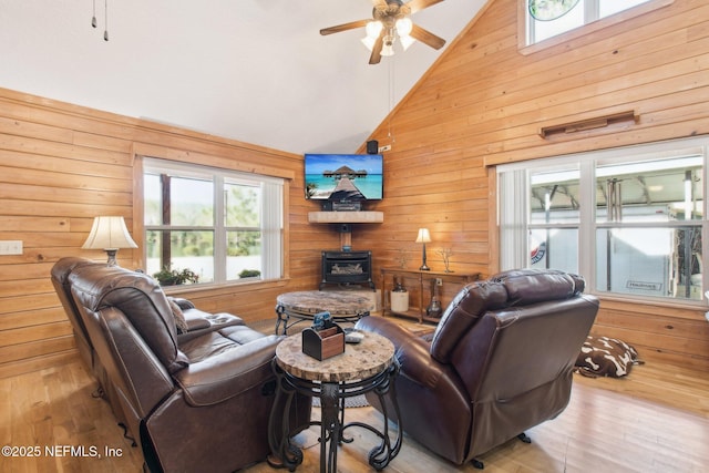 living room with high vaulted ceiling, wood walls, a wood stove, ceiling fan, and light hardwood / wood-style floors