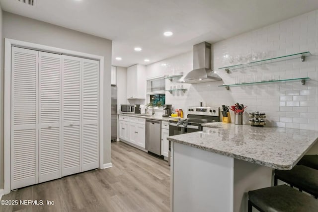 kitchen featuring white cabinetry, a kitchen breakfast bar, kitchen peninsula, stainless steel appliances, and wall chimney range hood