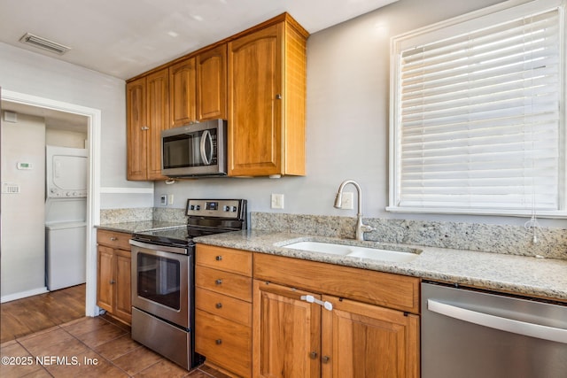 kitchen featuring visible vents, stacked washer and clothes dryer, appliances with stainless steel finishes, light stone countertops, and a sink