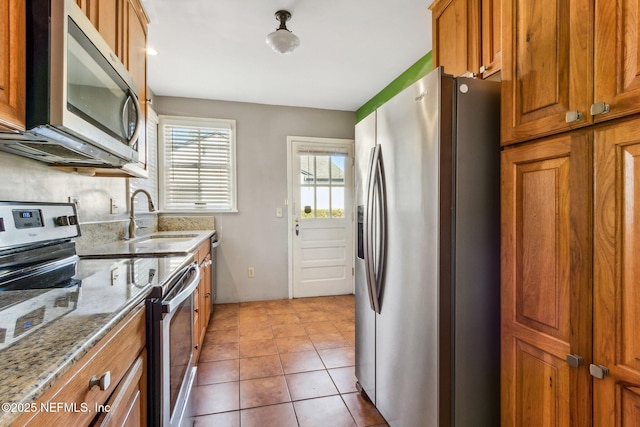 kitchen featuring light stone counters, appliances with stainless steel finishes, brown cabinetry, light tile patterned flooring, and a sink