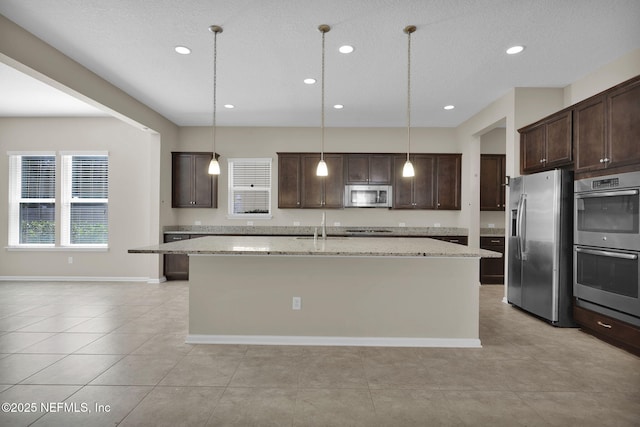 kitchen with dark brown cabinetry, light stone counters, decorative light fixtures, a center island with sink, and stainless steel appliances