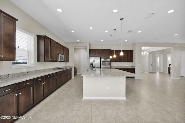 kitchen featuring dark brown cabinetry, light stone counters, hanging light fixtures, stainless steel appliances, and a kitchen island with sink