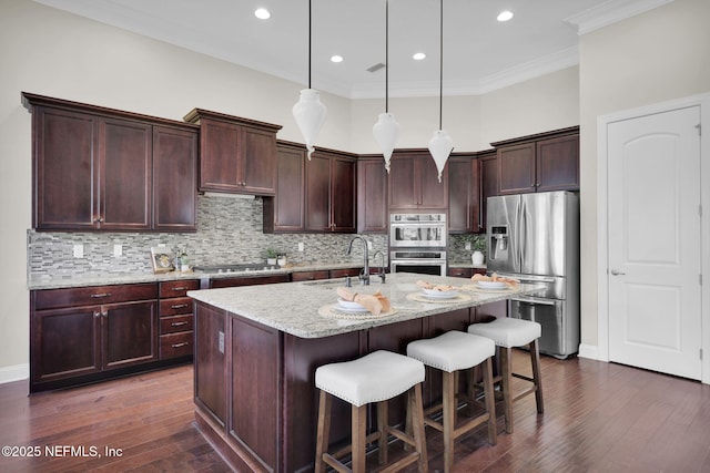 kitchen with sink, hanging light fixtures, stainless steel appliances, light stone counters, and an island with sink