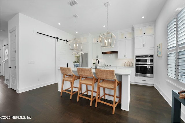 kitchen with decorative light fixtures, dark hardwood / wood-style floors, an island with sink, stainless steel double oven, and a barn door