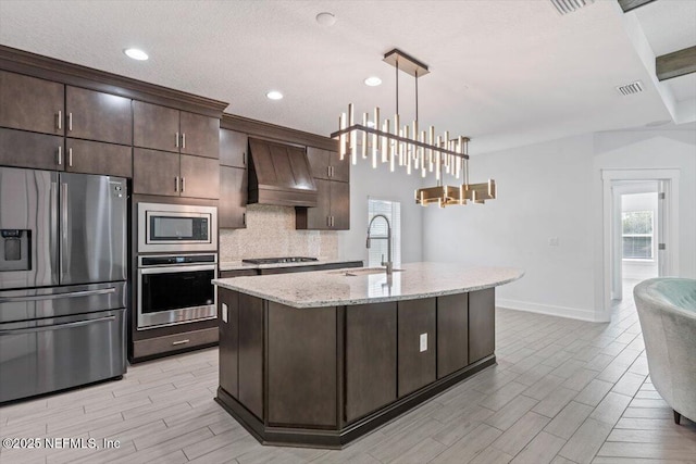 kitchen with an island with sink, stainless steel appliances, dark brown cabinetry, and premium range hood