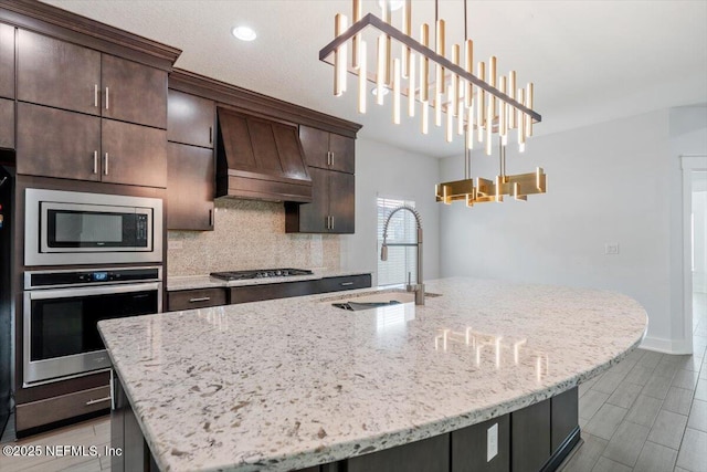 kitchen featuring appliances with stainless steel finishes, dark brown cabinetry, custom range hood, sink, and a kitchen island with sink