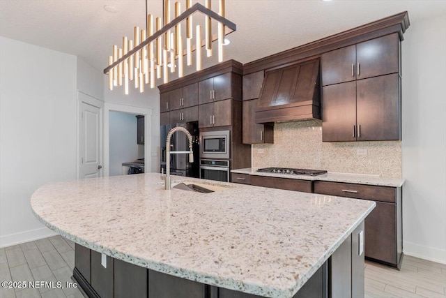 kitchen with a center island with sink, appliances with stainless steel finishes, dark brown cabinets, pendant lighting, and custom exhaust hood