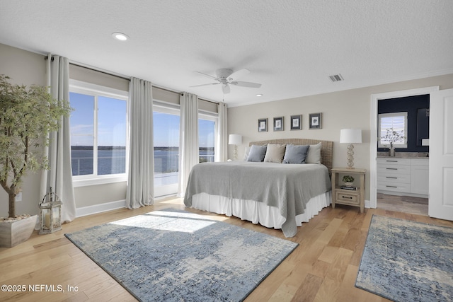 bedroom with light wood-type flooring, ensuite bathroom, a textured ceiling, and a water view