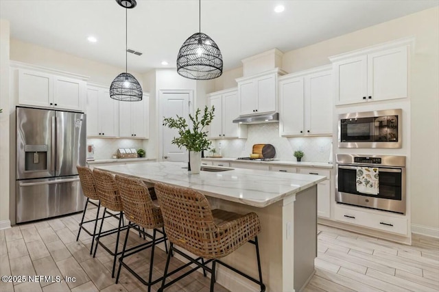 kitchen with light stone counters, an island with sink, pendant lighting, stainless steel appliances, and white cabinets