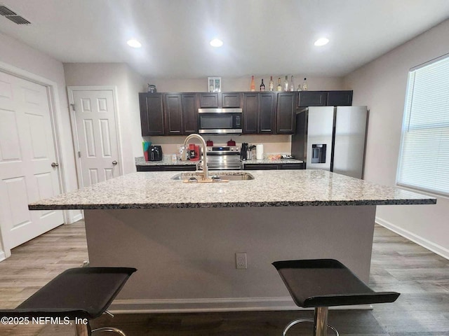 kitchen featuring dark brown cabinetry, light stone counters, light wood-type flooring, appliances with stainless steel finishes, and an island with sink