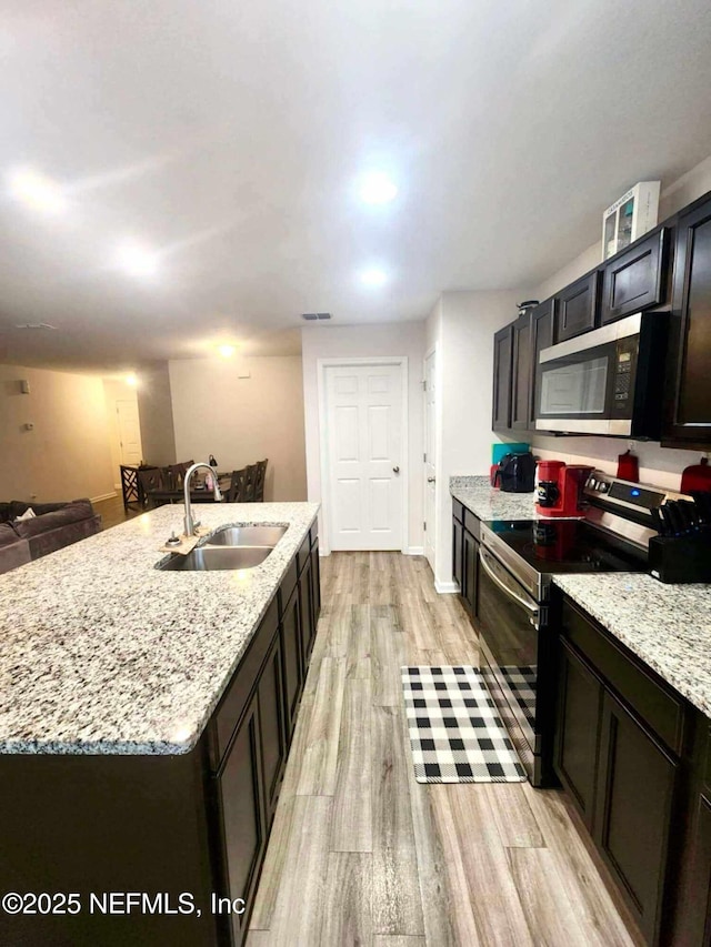 kitchen with a center island with sink, a sink, appliances with stainless steel finishes, light wood finished floors, and light stone countertops