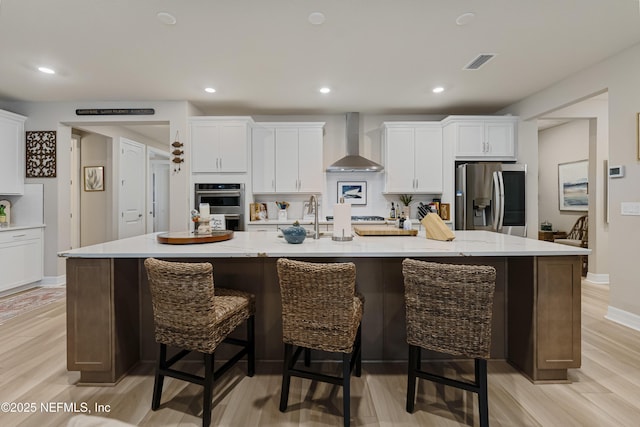 kitchen with wall chimney exhaust hood, a breakfast bar, a large island with sink, appliances with stainless steel finishes, and white cabinets