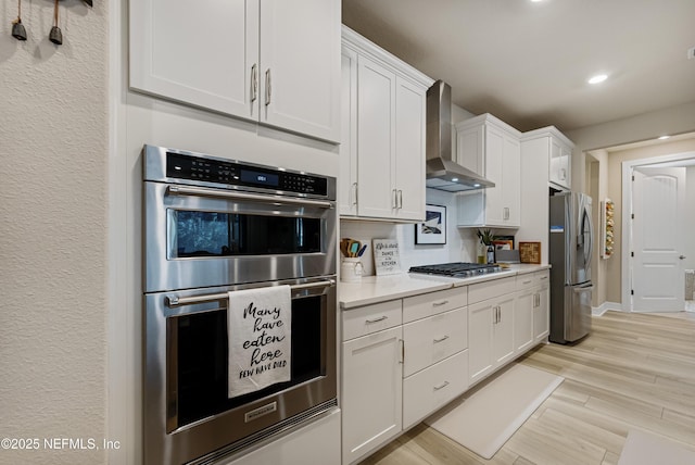 kitchen featuring white cabinetry, decorative backsplash, stainless steel appliances, wall chimney range hood, and light wood-type flooring