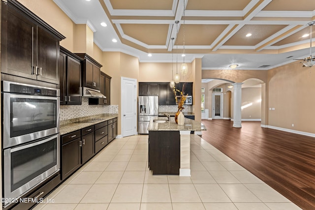 kitchen featuring dark brown cabinets, stainless steel appliances, decorative columns, light stone counters, and an island with sink