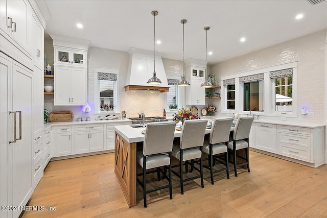 kitchen featuring a breakfast bar area, white cabinetry, an island with sink, custom range hood, and pendant lighting