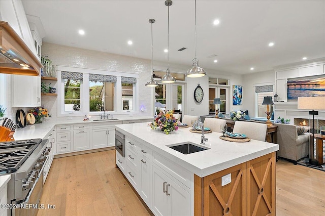 kitchen featuring custom exhaust hood, white cabinetry, hanging light fixtures, appliances with stainless steel finishes, and an island with sink