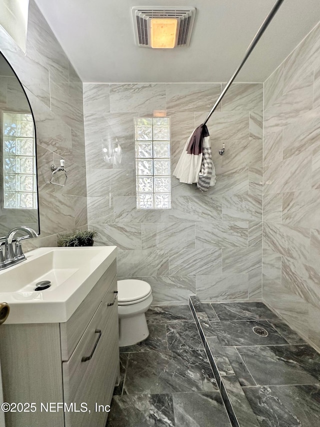 bathroom with a wealth of natural light, tile walls, vanity, tiled shower, and toilet