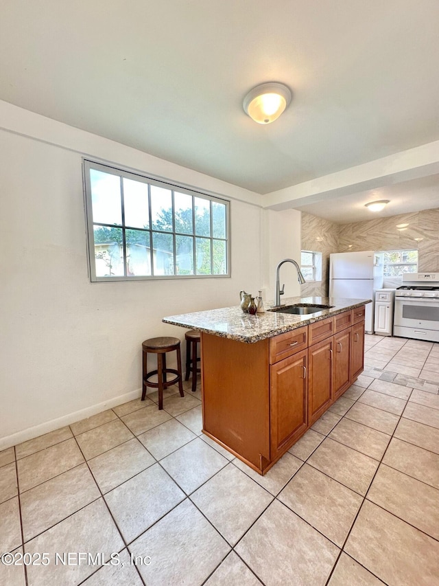 kitchen with light tile patterned flooring, sink, light stone counters, a center island with sink, and white appliances