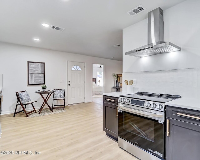 kitchen with wall chimney range hood, electric range, light hardwood / wood-style floors, and backsplash