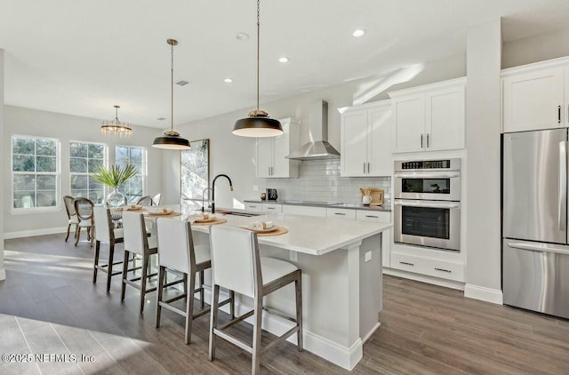 kitchen with appliances with stainless steel finishes, pendant lighting, an island with sink, white cabinets, and wall chimney range hood