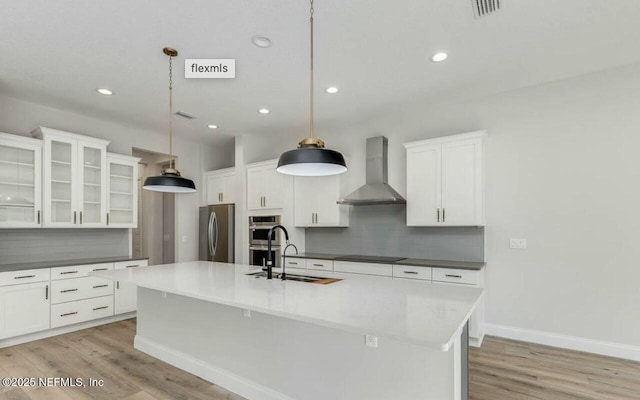 kitchen featuring wall chimney range hood, sink, appliances with stainless steel finishes, an island with sink, and decorative light fixtures