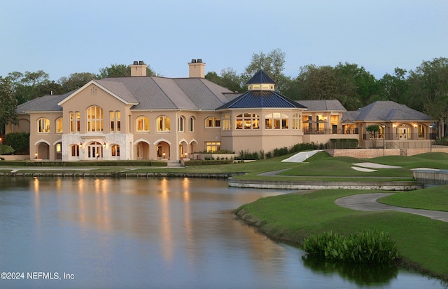 back house at dusk with a balcony, a water view, and a lawn