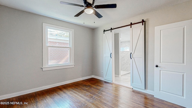 unfurnished bedroom with ceiling fan, ensuite bath, wood-type flooring, and a barn door