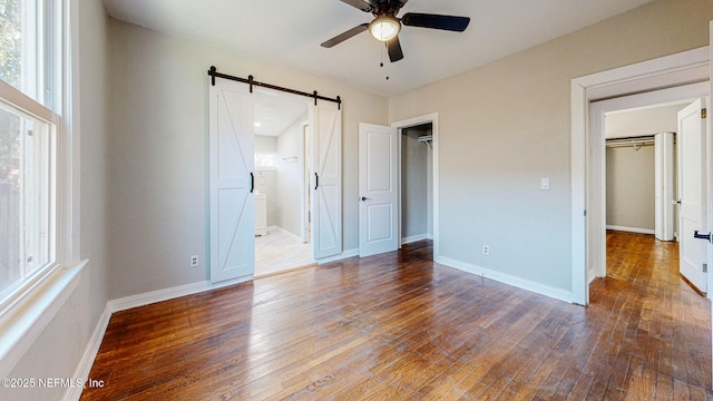 unfurnished bedroom with ensuite bath, dark wood-type flooring, a barn door, and ceiling fan