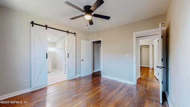 unfurnished bedroom featuring a walk in closet, ceiling fan, a barn door, dark wood-type flooring, and ensuite bath