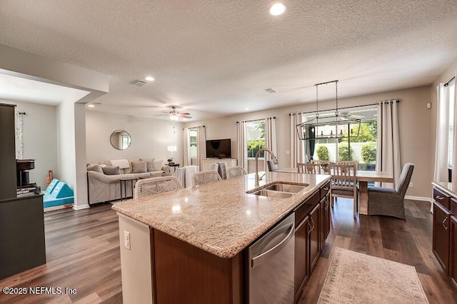 kitchen featuring sink, hanging light fixtures, stainless steel dishwasher, light stone counters, and a center island with sink