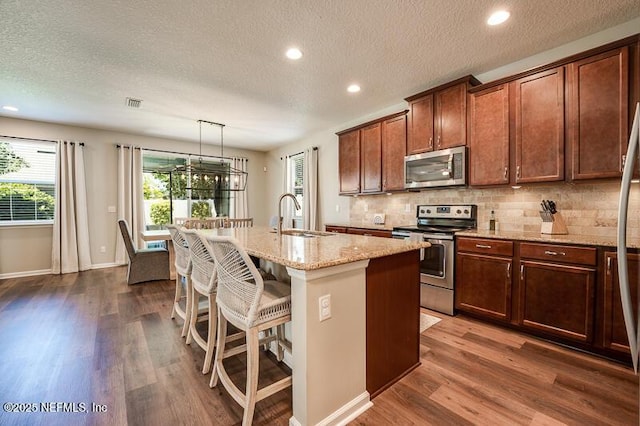 kitchen with dark wood-type flooring, stainless steel appliances, tasteful backsplash, an island with sink, and decorative light fixtures