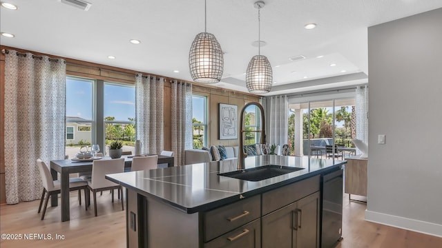 kitchen with an island with sink, dishwasher, sink, hanging light fixtures, and light wood-type flooring