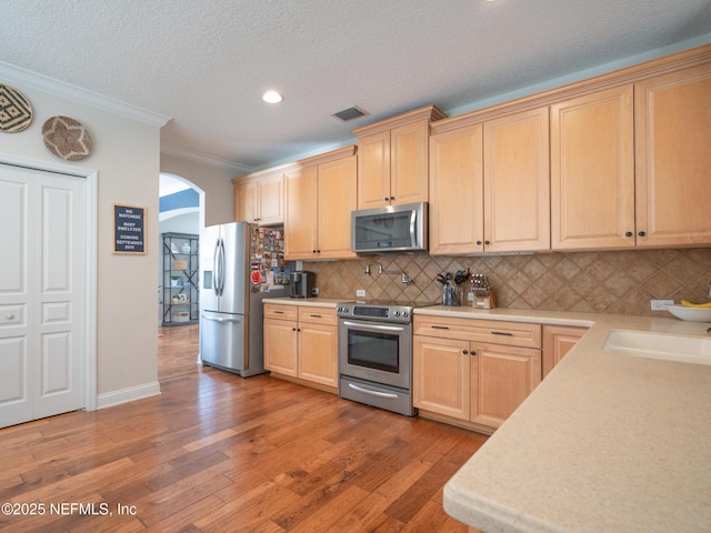 kitchen with appliances with stainless steel finishes, light brown cabinetry, backsplash, crown molding, and light hardwood / wood-style flooring